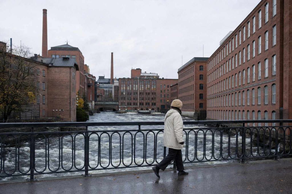 A walker crossing a bridge over the Tammerkoski rapids with old red-brick factory buildings behind